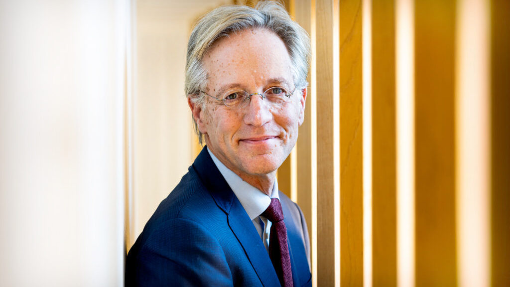 A smiling older man in a blue suit and glasses stands between wooden beams, looking at the camera in a brightly lit space.