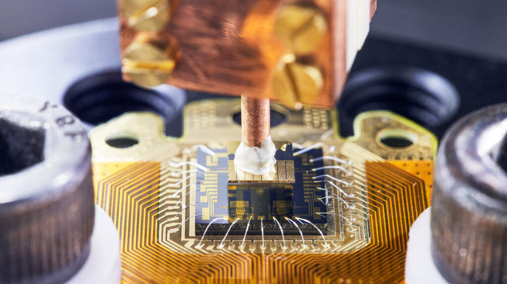 Close-up of a quantum computer chip with wires attached and a metal probe positioned above, showing intricate circuitry and components.