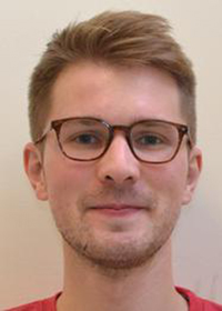 A young man with light skin, short brown hair, and glasses is facing the camera and smiling slightly. He is wearing a red shirt and is in front of a plain, light-colored background.