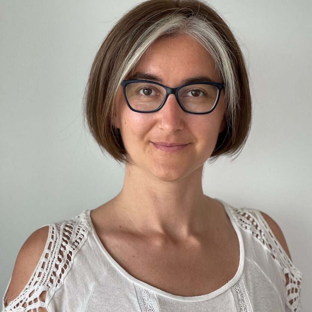 A woman with short brown hair and a streak of white, wearing glasses and a white top with lace details, stands in front of a plain light background, smiling softly at the camera.
