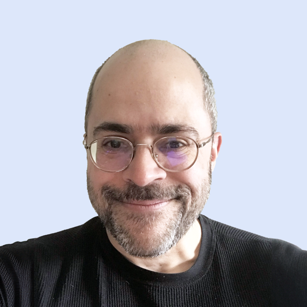 A smiling man with glasses, a beard, and a mustache, wearing a black sweater in front of a plain light blue background.