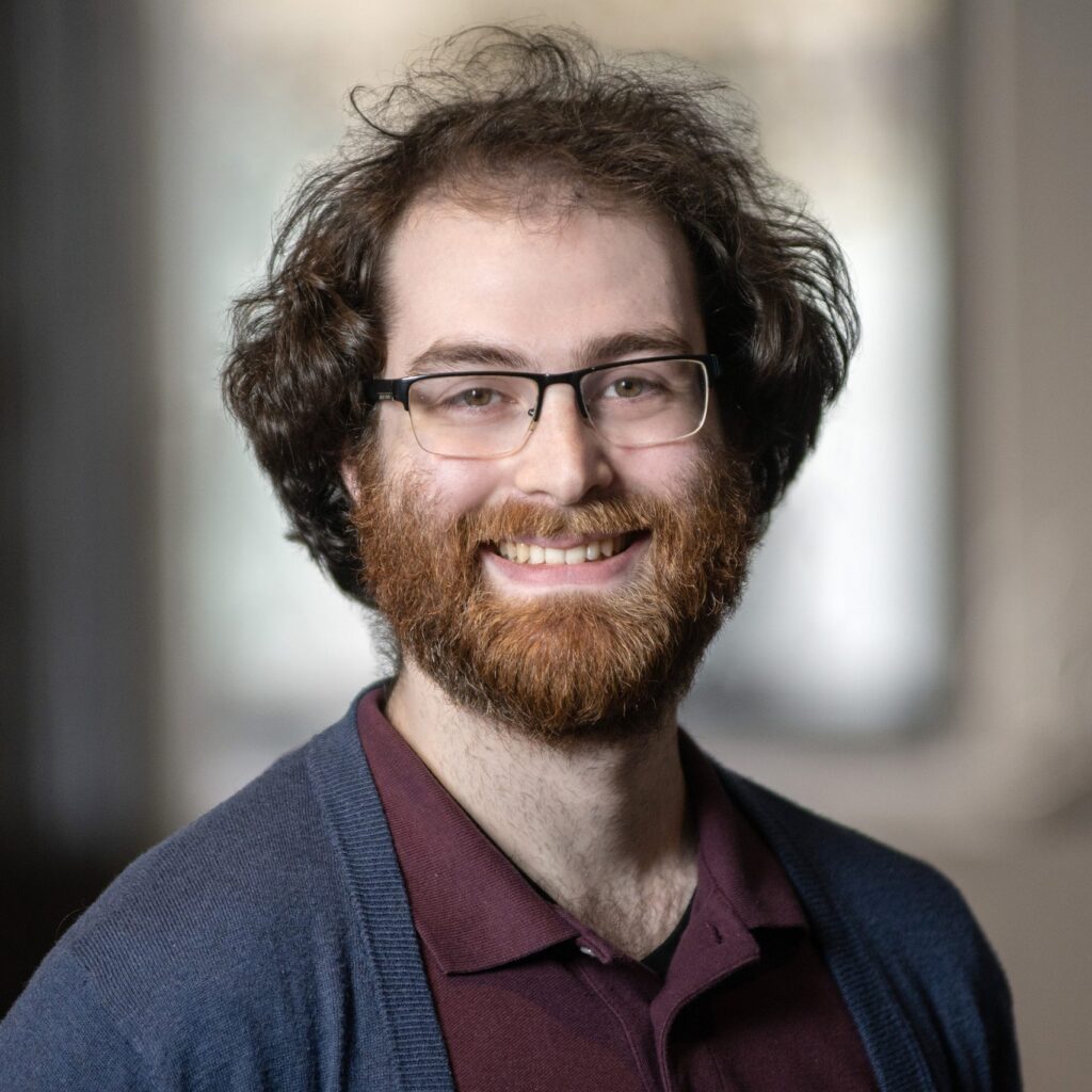 A man with curly brown hair, a full beard, and glasses smiles at the camera. He is wearing a maroon shirt and a blue cardigan, with a blurred indoor background.