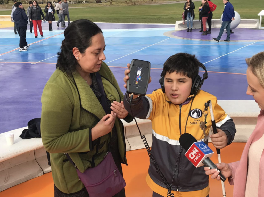 A boy wearing headphones holds up an audio device while a woman stands beside him and another person holds a CNN Chile microphone toward him. People are in the colorful outdoor area in the background.
