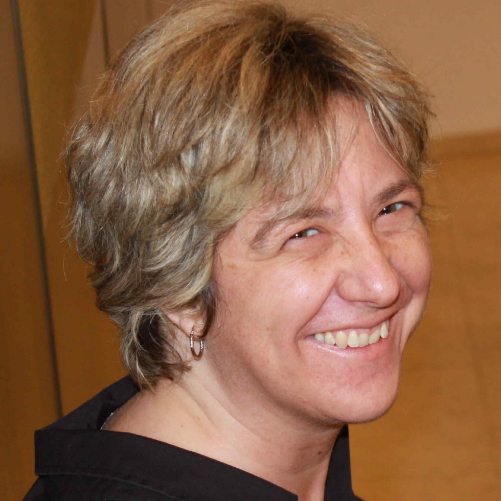 A person with short, light brown hair is smiling and looking over their shoulder. They are wearing a black top and a small hoop earring. The background is softly blurred.