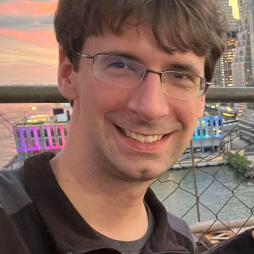 A smiling man with glasses and brown hair stands in front of a cityscape at sunset, wearing a black Iowa State University jacket.