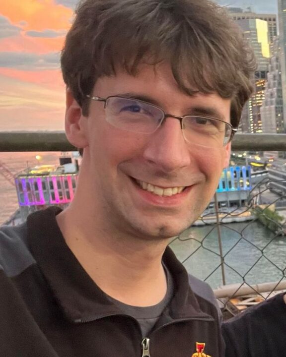 A smiling man with glasses and brown hair stands in front of a cityscape at sunset, wearing a black Iowa State University jacket.