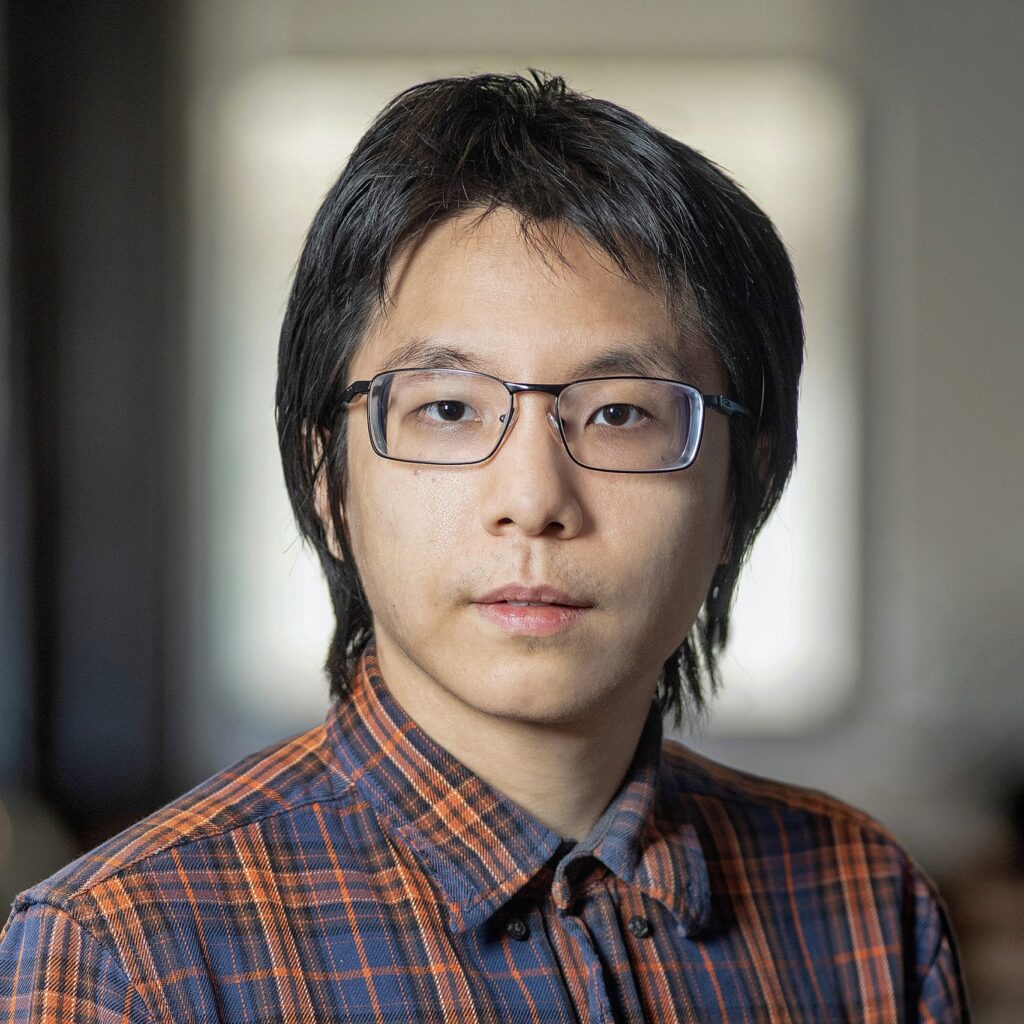 A person with dark hair and glasses wearing a plaid shirt looks at the camera with a neutral expression. The background is softly blurred, suggesting an indoor setting.