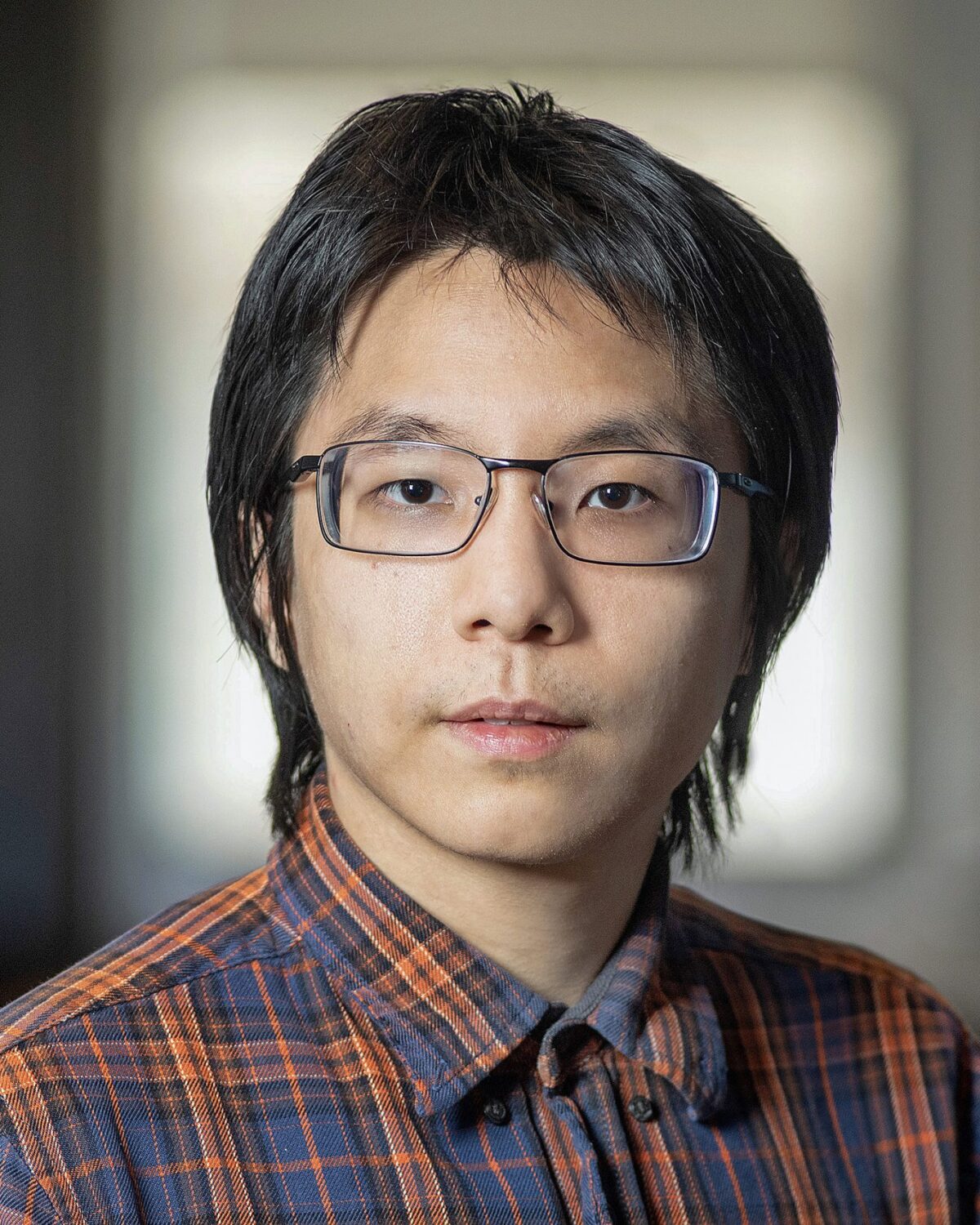 A person with dark hair and glasses wearing a plaid shirt looks at the camera with a neutral expression. The background is softly blurred, suggesting an indoor setting.