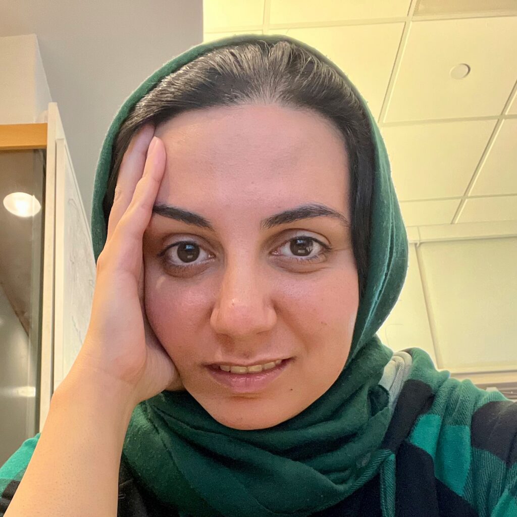 A person wearing a green headscarf rests their face on their hand, looking directly at the camera with a slight smile. The background shows a ceiling with fluorescent lights.