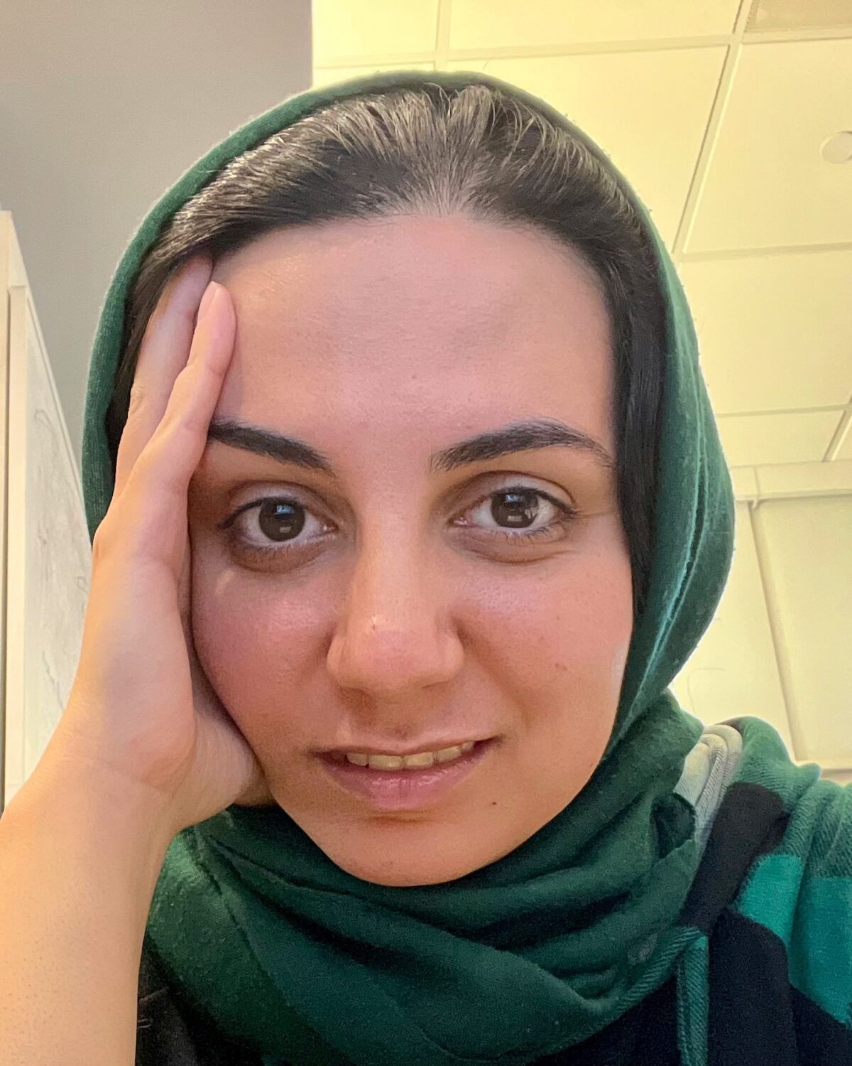 A person wearing a green headscarf rests their face on their hand, looking directly at the camera with a slight smile. The background shows a ceiling with fluorescent lights.