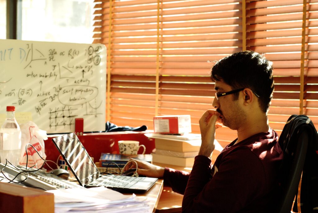 A person sits at a cluttered desk, looking thoughtful and using a laptop. Sunlight streams through wooden blinds. A whiteboard with notes and diagrams is in the background.