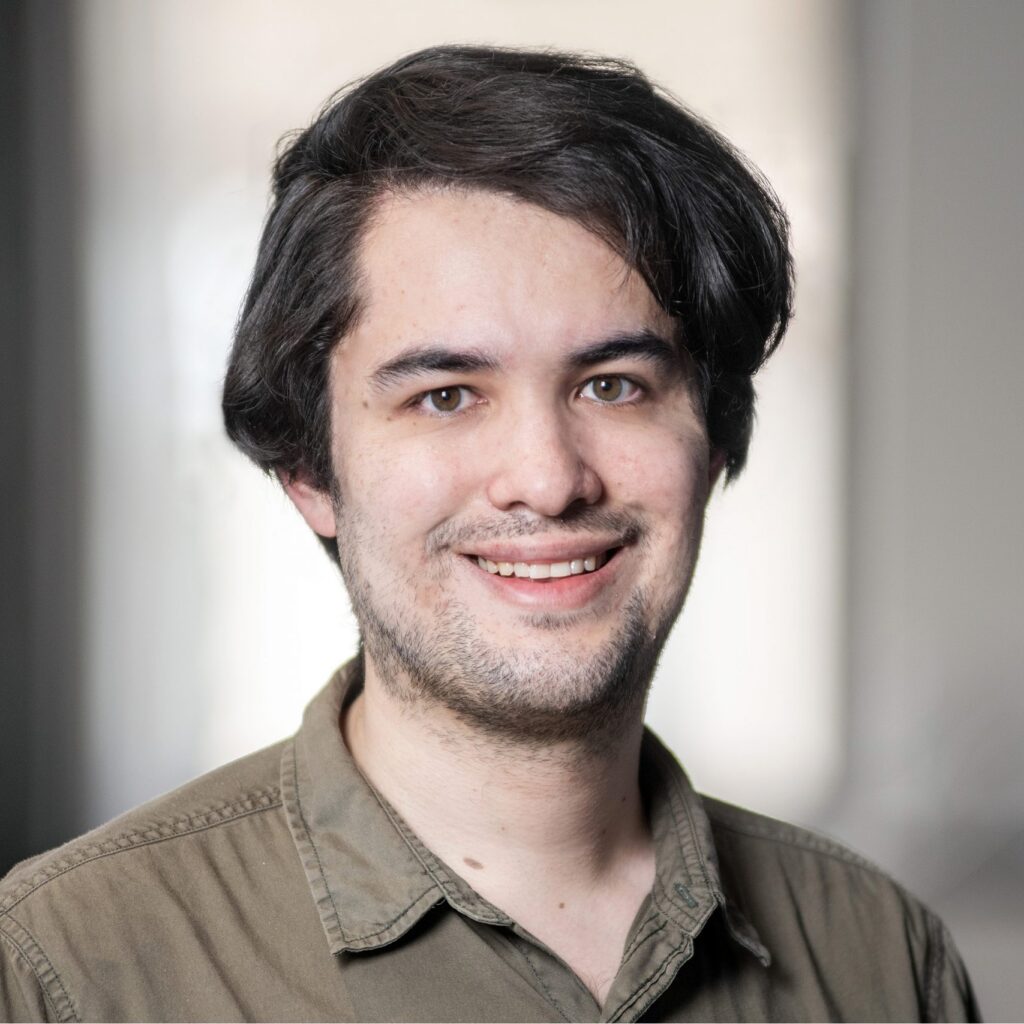 A young man with medium-length dark hair and a trimmed beard is smiling at the camera. He is wearing an olive-green button-up shirt. The background is softly blurred indoors.