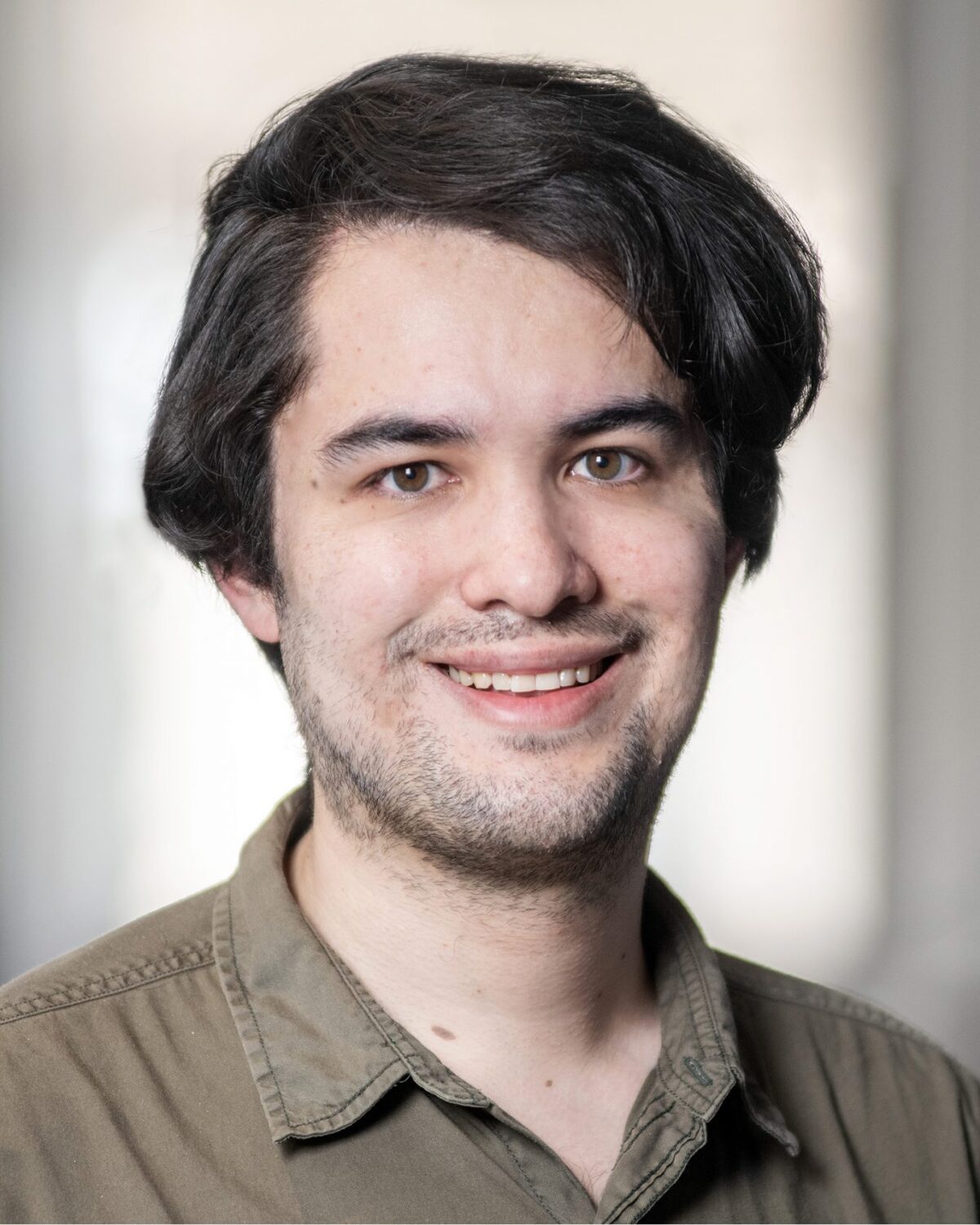 A young man with medium-length dark hair and a trimmed beard is smiling at the camera. He is wearing an olive-green button-up shirt. The background is softly blurred indoors.