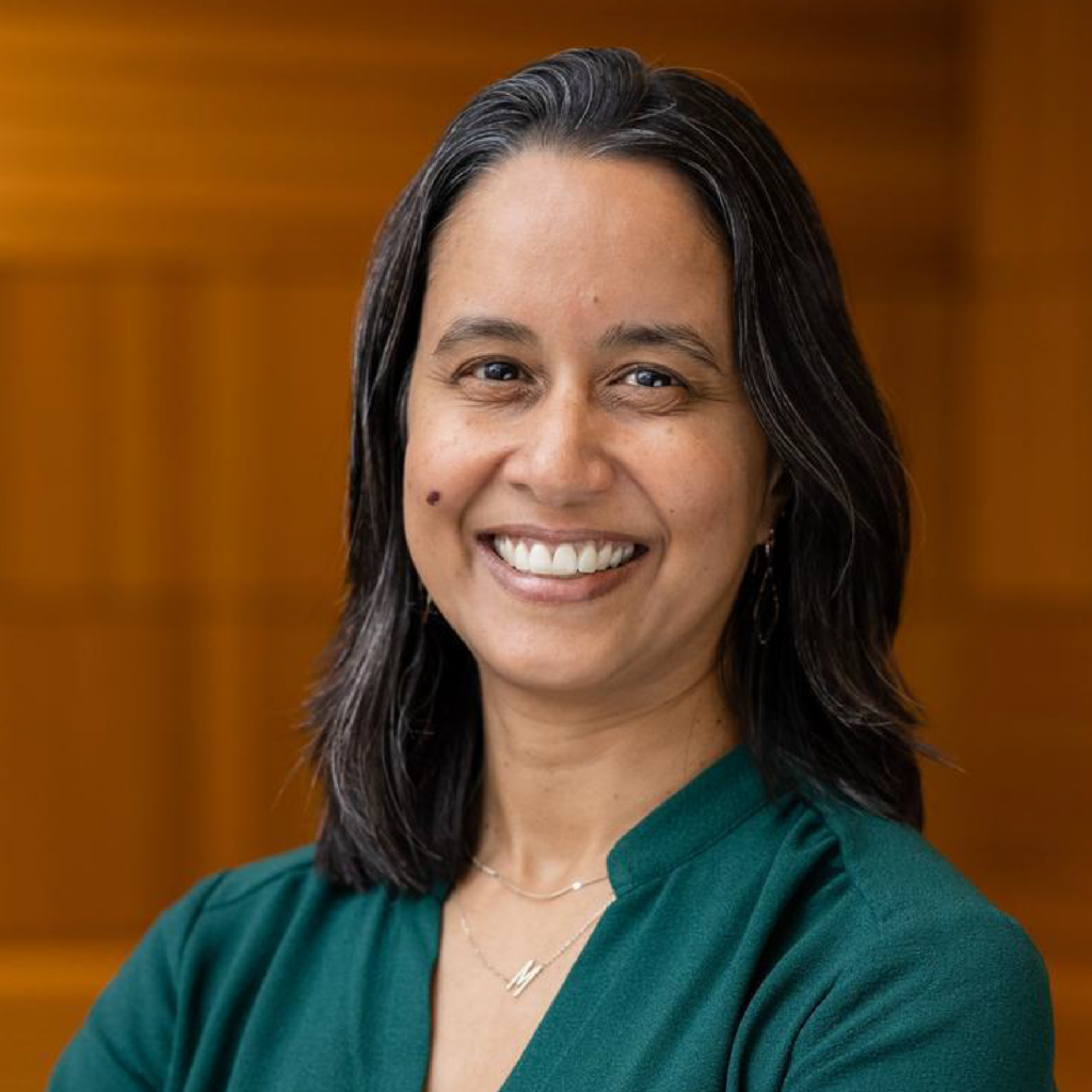 A woman with long dark hair and a green top smiles at the camera, standing in front of a blurred, warm-toned background.