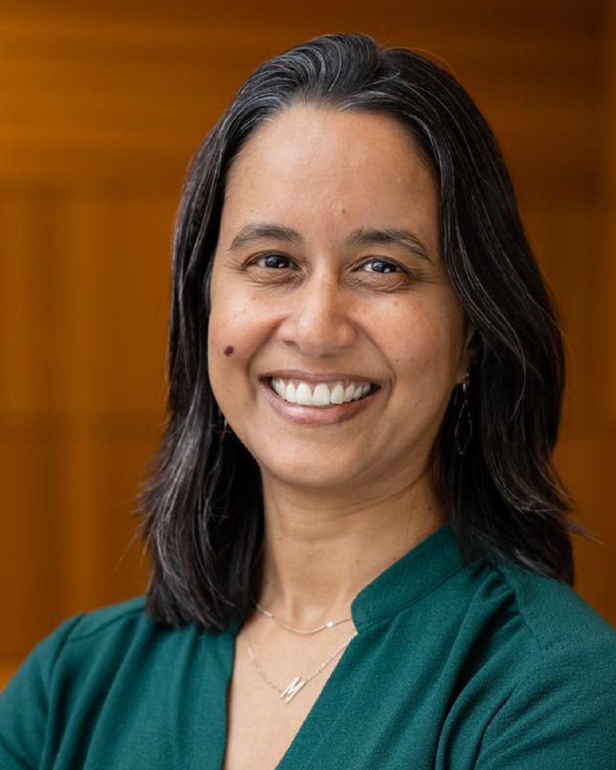 A woman with long dark hair and a green top smiles at the camera, standing in front of a blurred, warm-toned background.