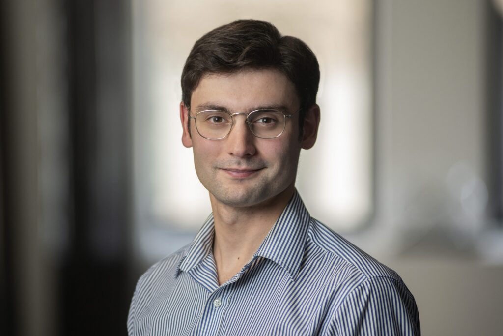 A young man with short brown hair and glasses, wearing a blue striped shirt, stands indoors in front of a blurred background, looking at the camera with a slight smile.