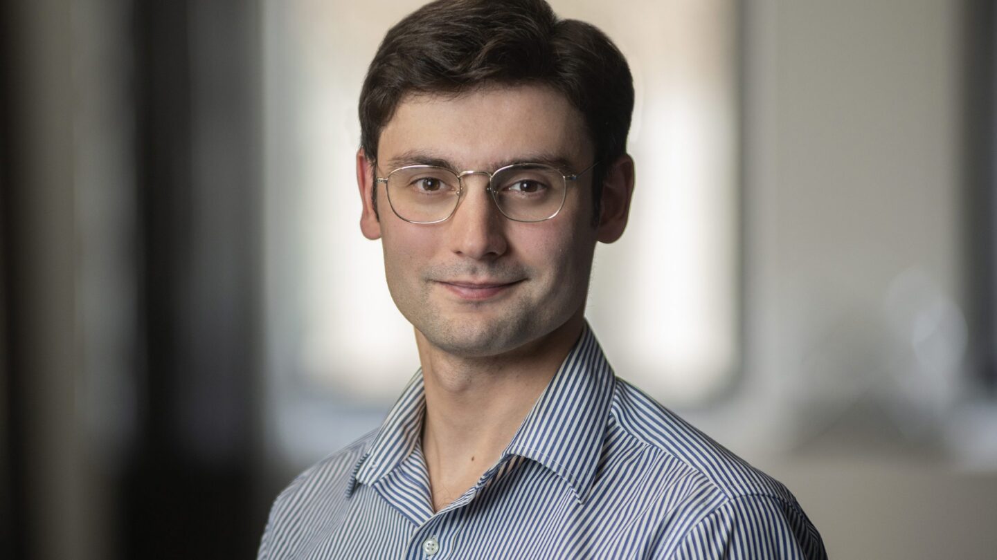 A young man with short brown hair and glasses, wearing a blue striped shirt, stands indoors in front of a blurred background, looking at the camera with a slight smile.
