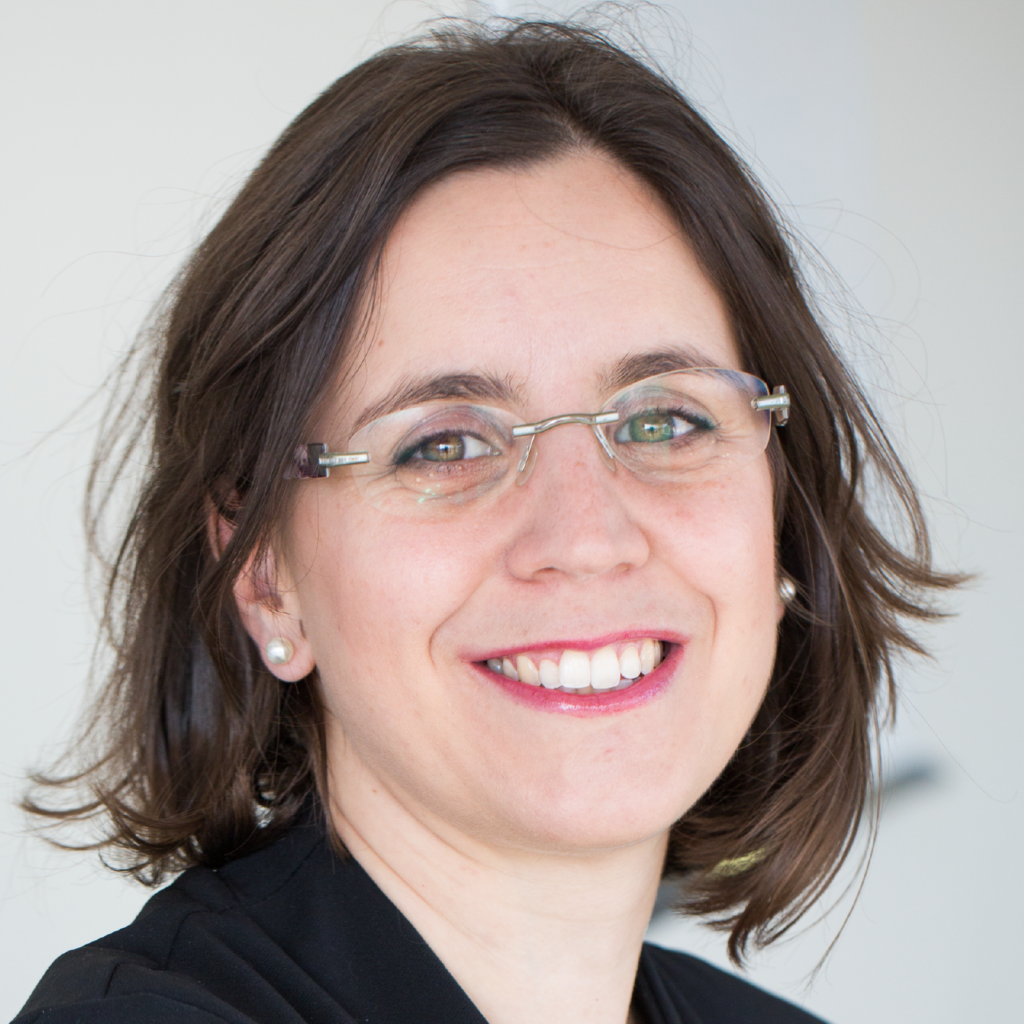 Woman with shoulder-length brown hair, wearing glasses and a black top, smiling at the camera in a bright indoor setting.