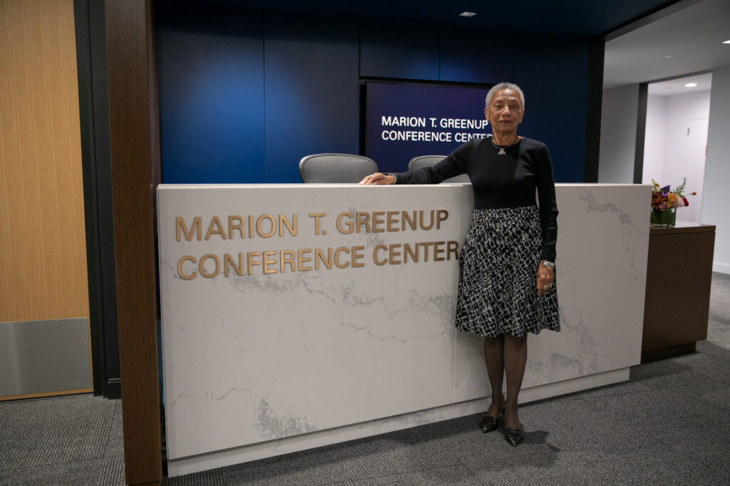 A woman stands beside a white reception desk labeled Marion T. Greenup Conference Center in a modern office lobby. She is wearing a black top and patterned skirt.