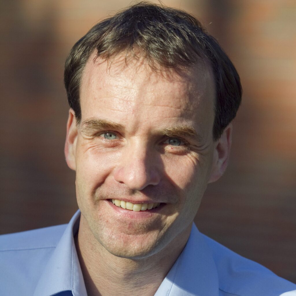 A man with short brown hair and blue eyes smiles outdoors, wearing a light blue collared shirt. The background is softly blurred with warm light and hints of a brick wall.