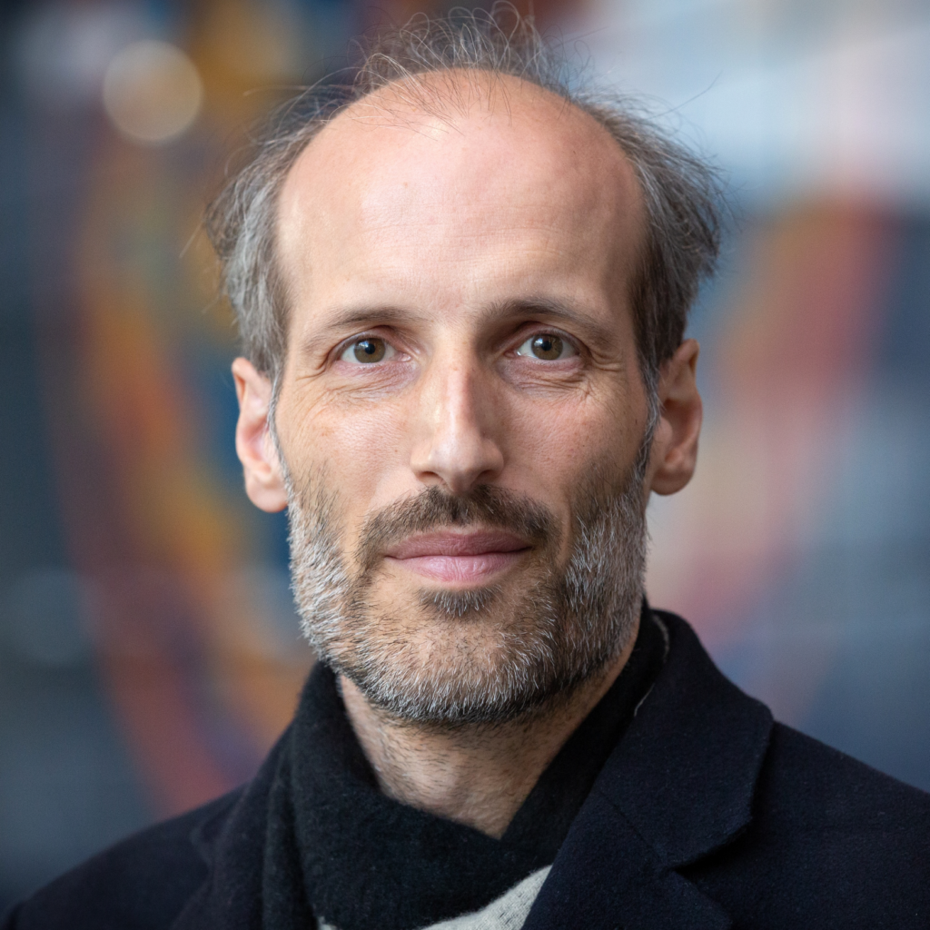 A middle-aged man with a receding hairline and gray beard looks at the camera, wearing a dark coat and black scarf, with a blurred, colorful background.