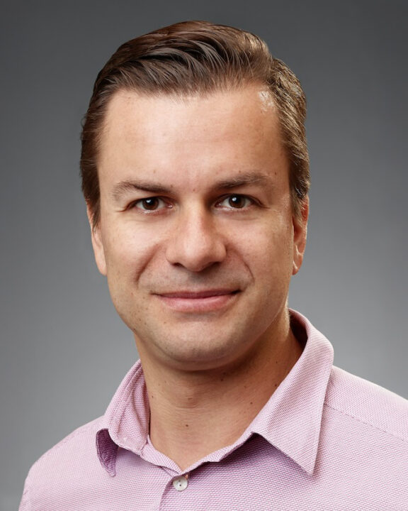 A man with short brown hair wearing a light pink collared shirt smiles at the camera against a plain gray background.