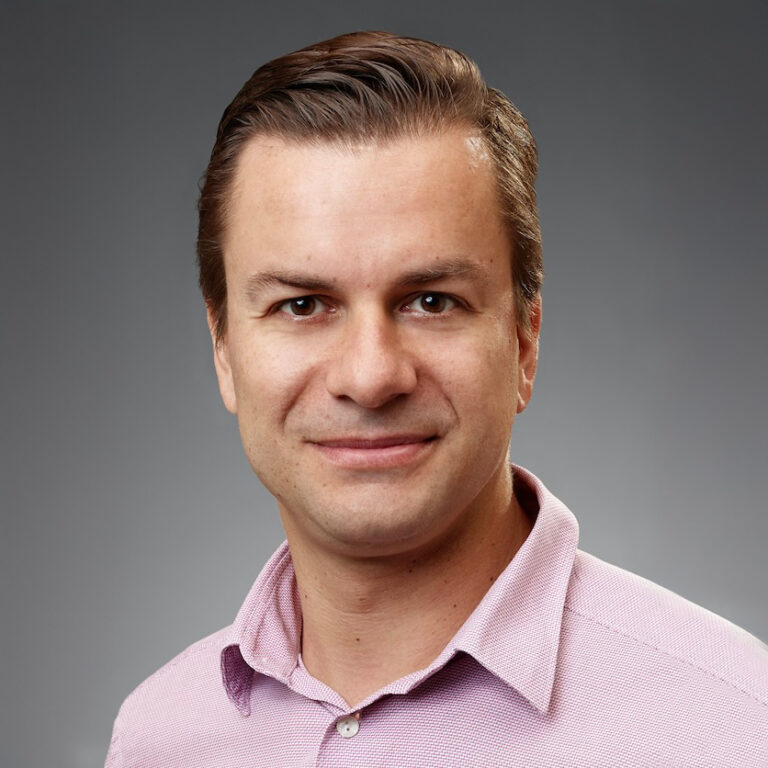 A man with short brown hair wearing a light pink collared shirt smiles at the camera against a plain gray background.