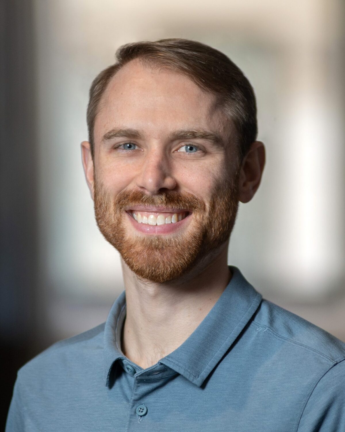 A young man with short brown hair and a beard, smiling, wearing a light blue collared shirt, standing indoors with a blurred background.