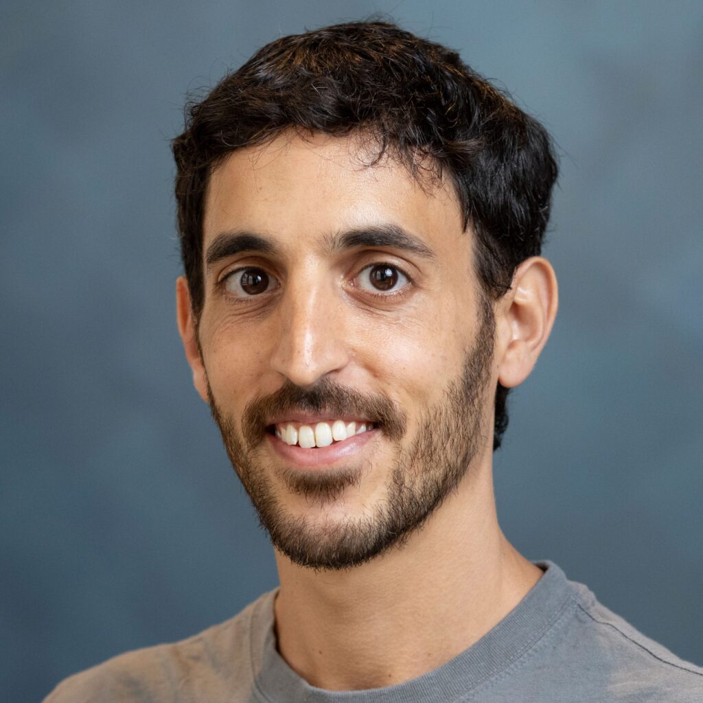 A man with short dark hair, beard, and mustache, wearing a grey shirt, smiling at the camera with a blue-gray background.