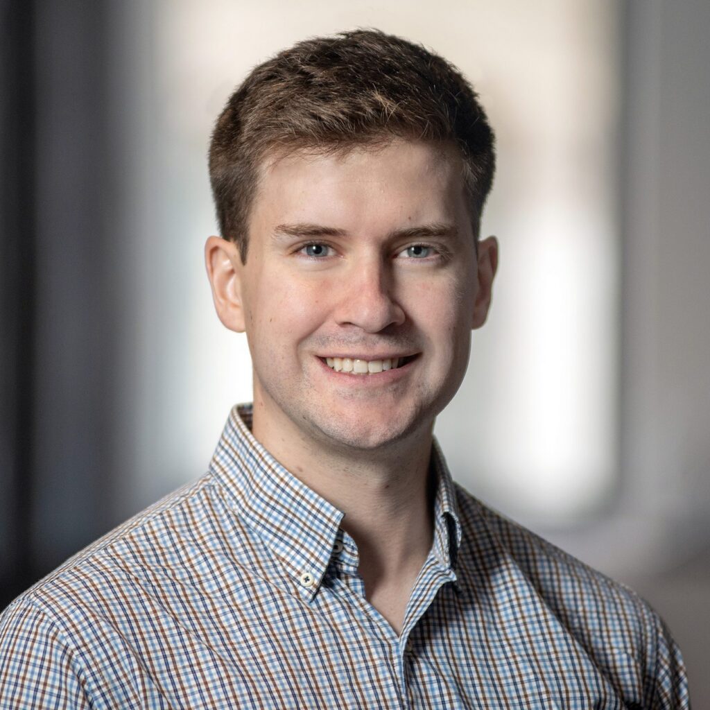 A young man with short brown hair, wearing a checked button-up shirt, smiles at the camera. The background is softly blurred with neutral colors.