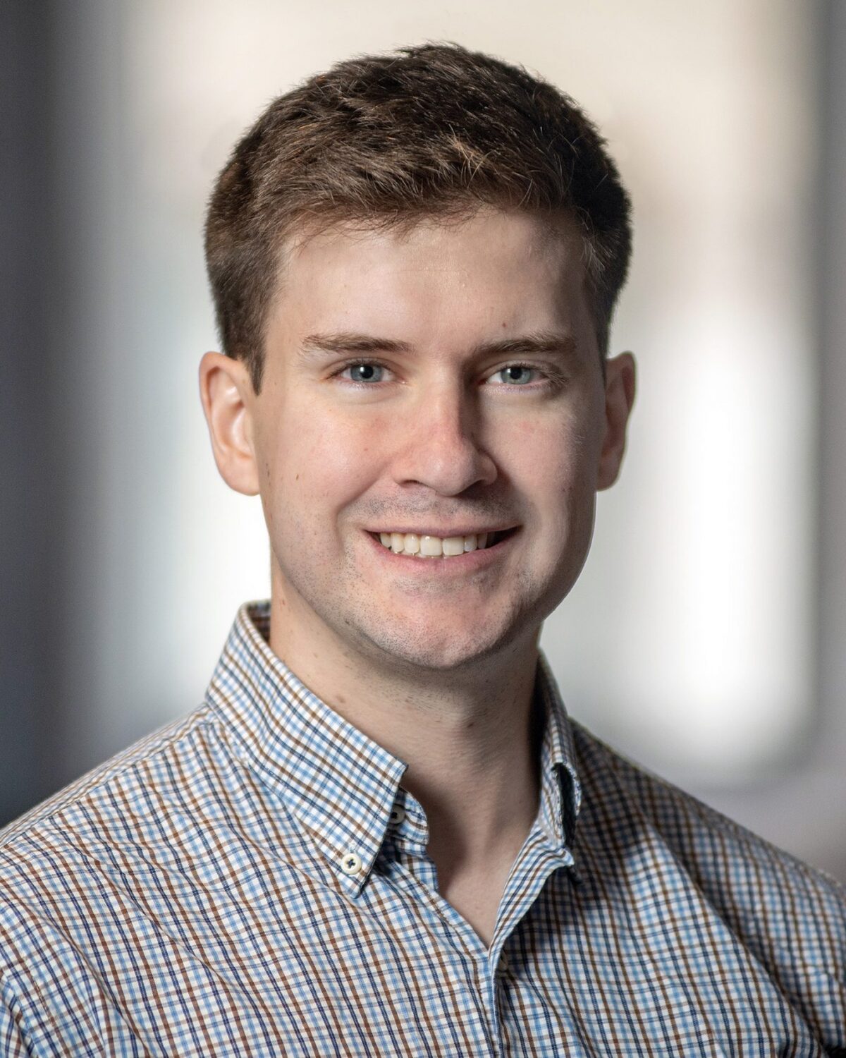 A young man with short brown hair, wearing a checked button-up shirt, smiles at the camera. The background is softly blurred with neutral colors.