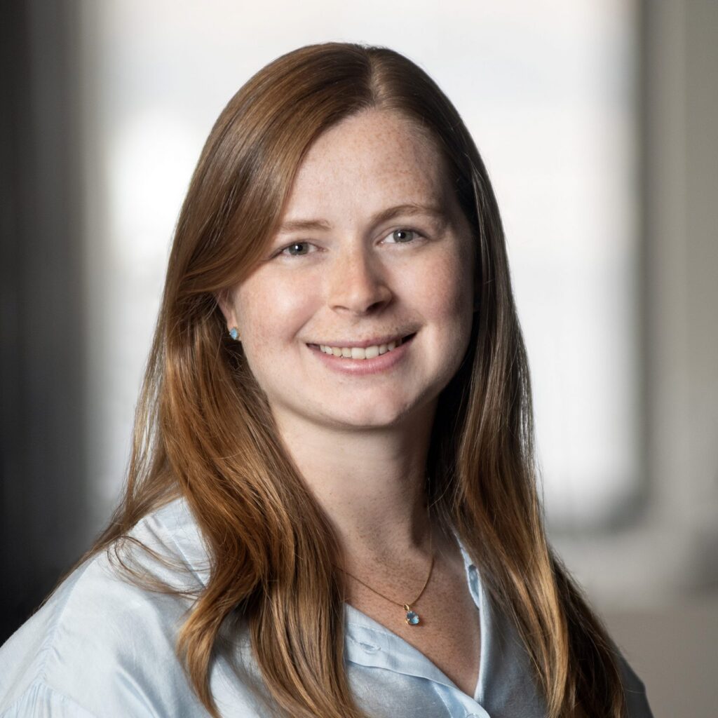 A woman with long red hair, wearing a light blue blouse and a necklace, smiles at the camera in a softly lit indoor setting with a blurred background.