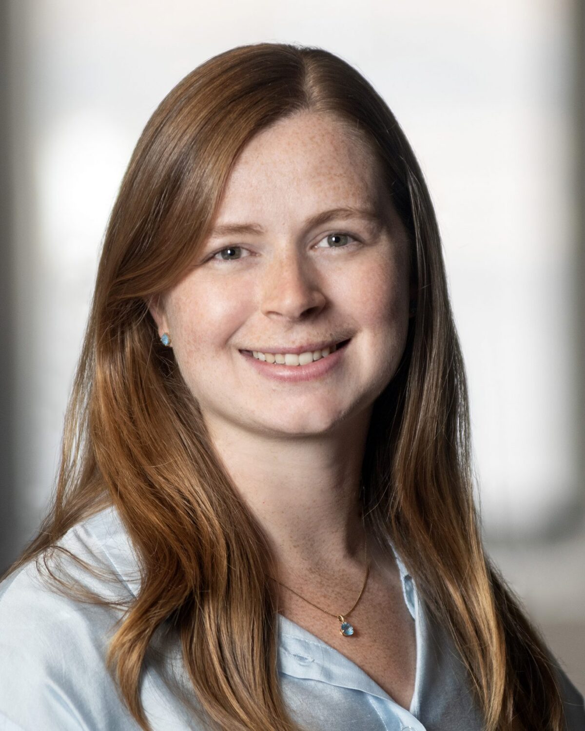 A woman with long red hair, wearing a light blue blouse and a necklace, smiles at the camera in a softly lit indoor setting with a blurred background.