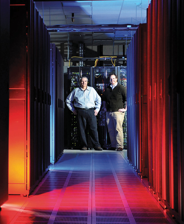 Two people stand in a brightly lit server room aisle, surrounded by tall racks of computer servers with red and blue lighting illuminating the floor and walls.