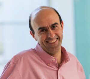 A man with a receding hairline, wearing a pink button-up shirt, smiles warmly at the camera. The background is softly blurred with light blue and white tones.