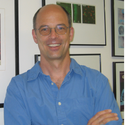 A person wearing glasses and a blue shirt stands smiling with arms crossed in front of a wall covered with framed pictures and images.
