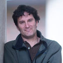 A man with dark curly hair, wearing a dark green jacket over a brown shirt, stands indoors leaning against a wall and smiling softly at the camera. The background is softly blurred.