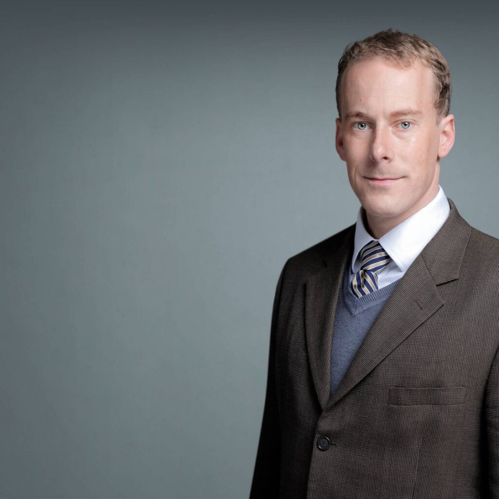 A man in a brown suit, blue sweater, and striped tie stands against a plain gray background, looking at the camera and smiling slightly.
