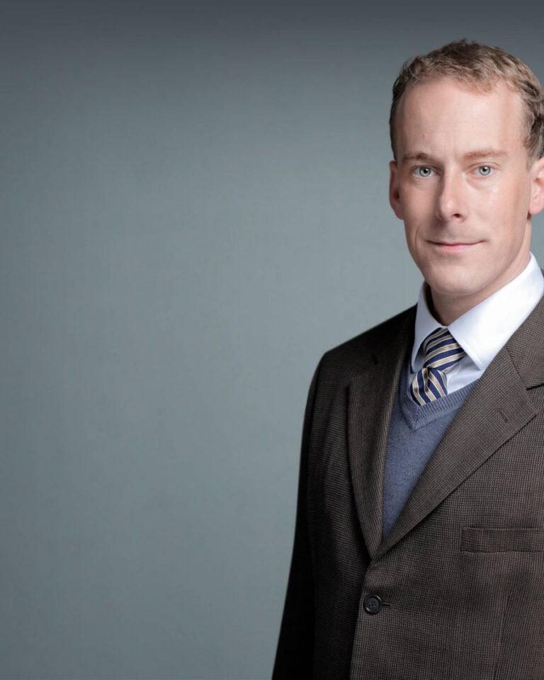 A man in a brown suit, blue sweater, and striped tie stands against a plain gray background, looking at the camera and smiling slightly.