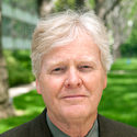 An older man with light gray hair wearing a dark suit stands outside on a sunny day, with green grass and trees in the blurred background.