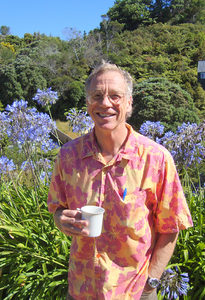 A man wearing a colorful floral shirt holds a white cup and smiles while standing outside among green plants and purple flowers, with trees and blue sky in the background.