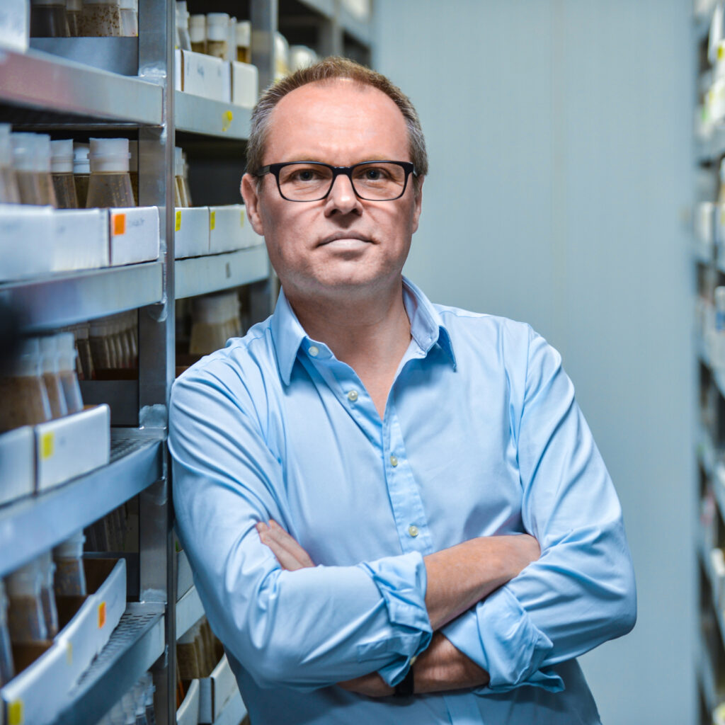 A man in glasses and a light blue shirt stands with arms crossed between shelves filled with jars in a storage or laboratory setting.
