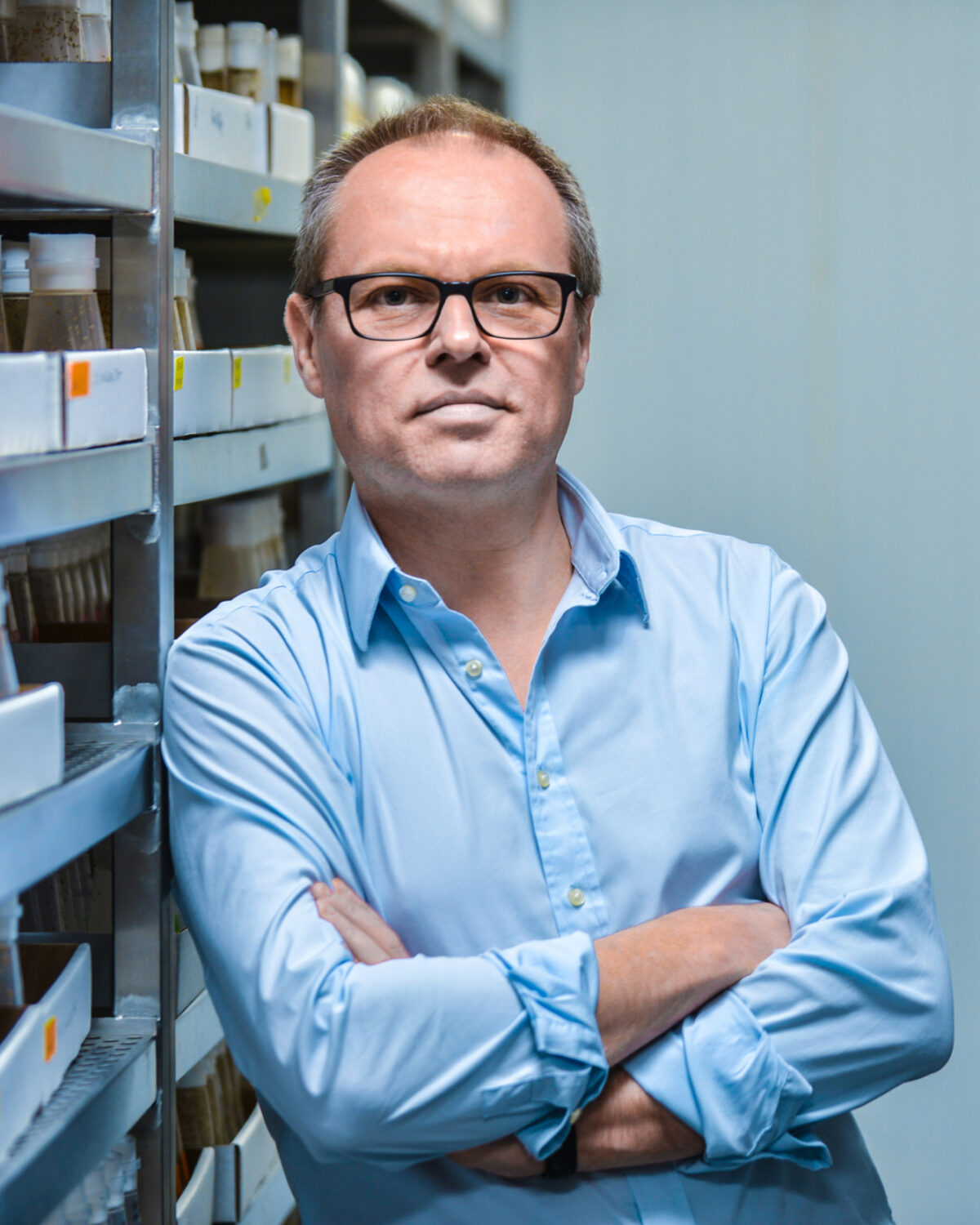A man in glasses and a light blue shirt stands with arms crossed between shelves filled with jars in a storage or laboratory setting.