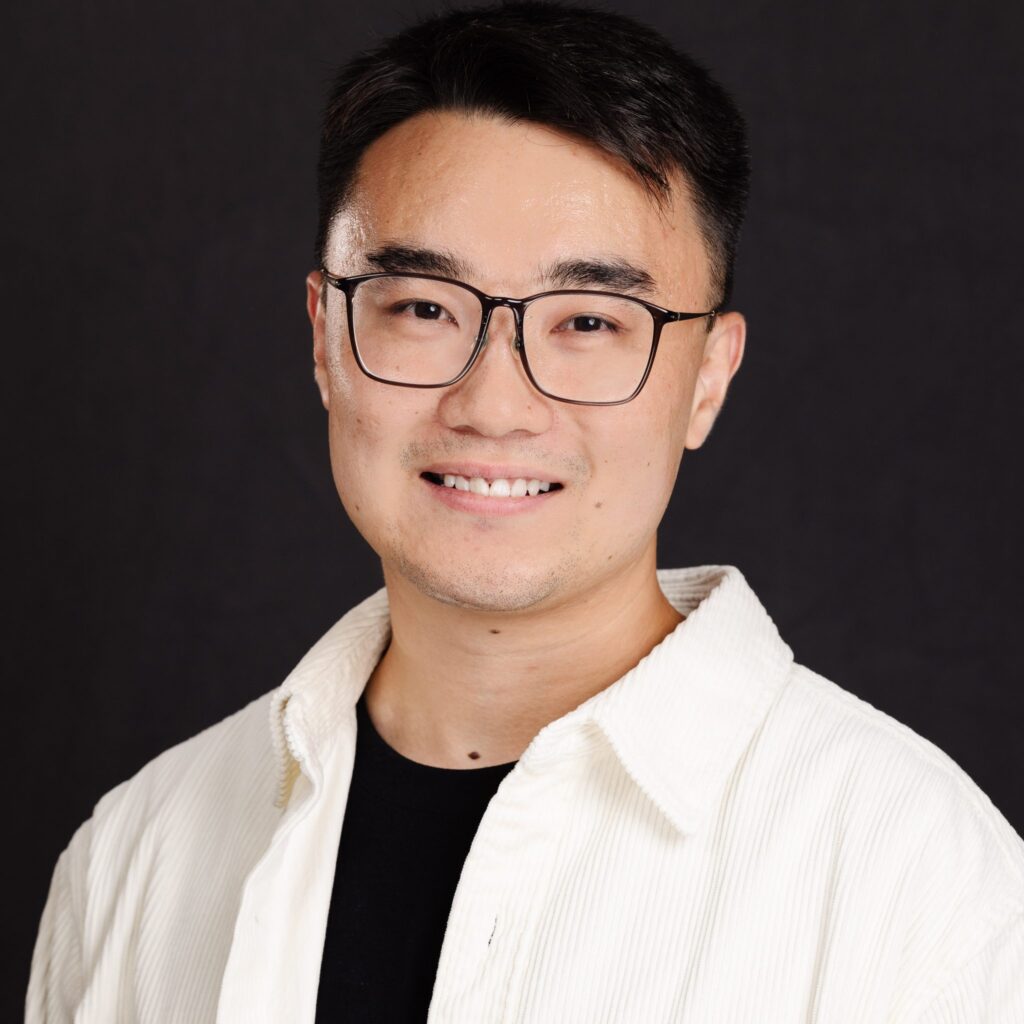 A person with short dark hair and glasses, wearing a white jacket over a black shirt, smiles at the camera against a plain dark background.