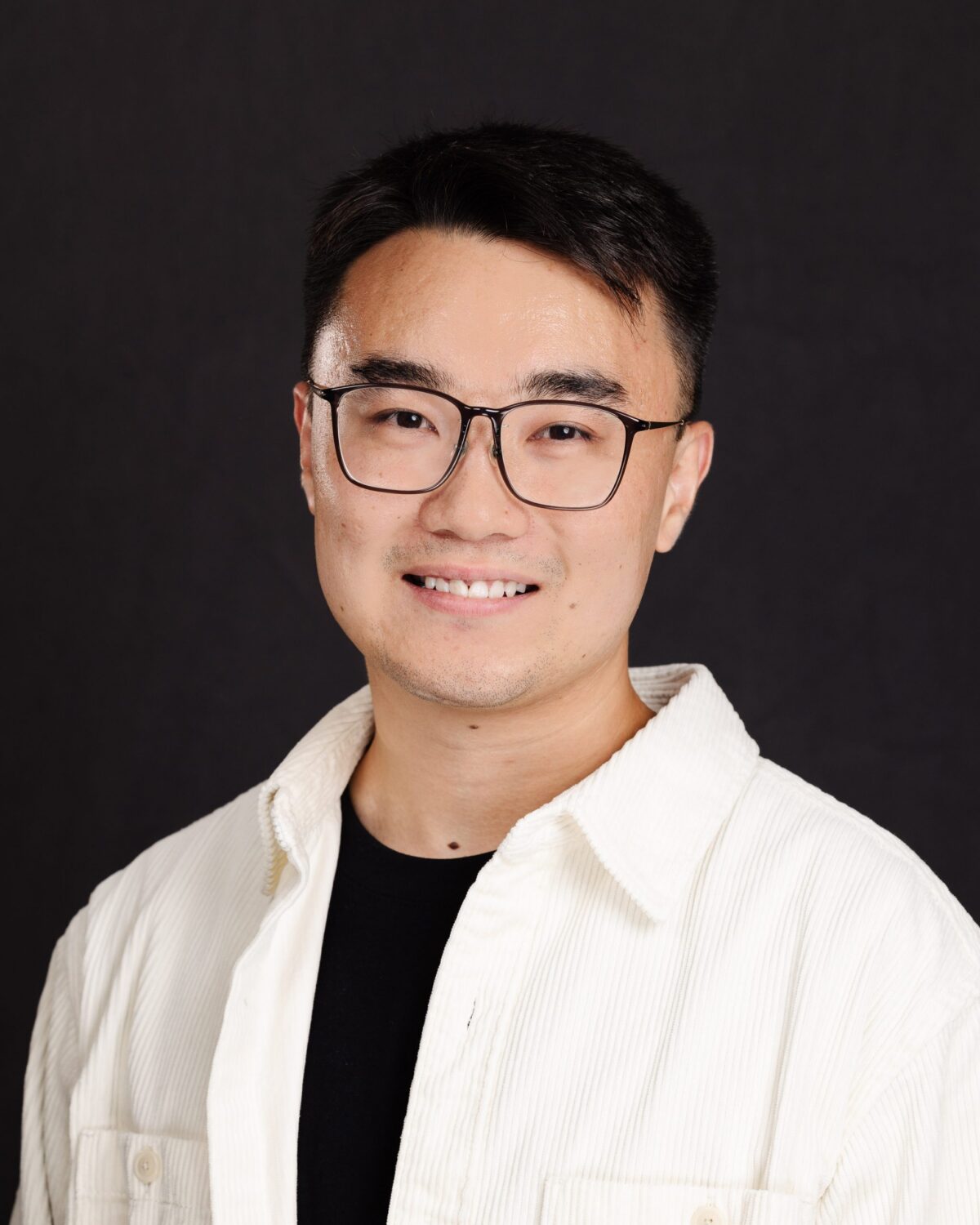 A person with short dark hair and glasses, wearing a white jacket over a black shirt, smiles at the camera against a plain dark background.