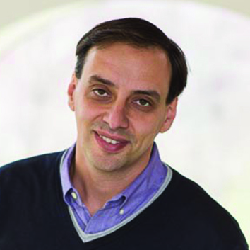 A man with short dark hair, wearing a blue collared shirt and a dark sweater, smiles slightly while posing in front of a light, blurred background.