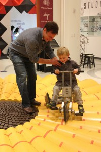 An adult helps a young boy ride a small pedal bike on a wavy yellow track inside a science or discovery museum.