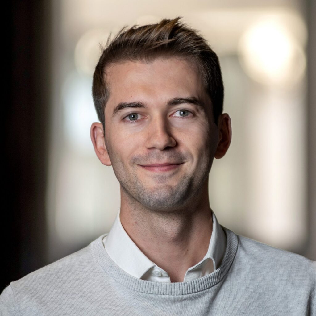 A man with short brown hair wearing a white collared shirt and light sweater smiles at the camera. The background is softly blurred, drawing focus to his face.