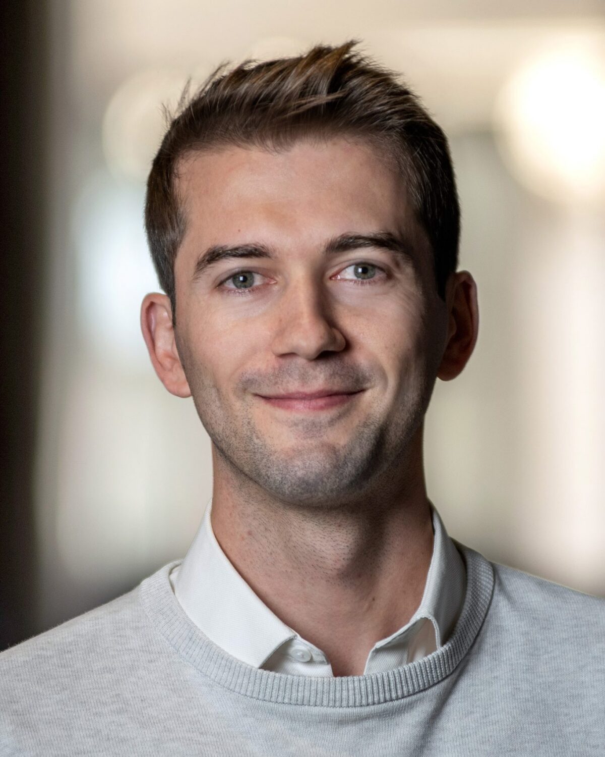 A man with short brown hair wearing a white collared shirt and light sweater smiles at the camera. The background is softly blurred, drawing focus to his face.