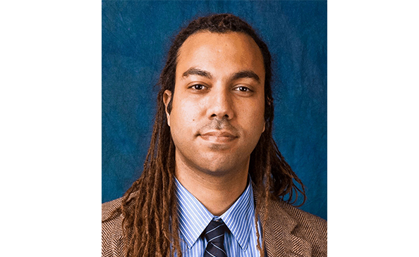 A man with long dreadlocks wearing a brown suit jacket, blue dress shirt, and striped tie, poses in front of a solid blue background.